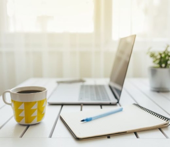 A laptop on a table with a notepad and a cup of coffee